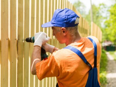 A picture of a man installing fence boards with a power drill.